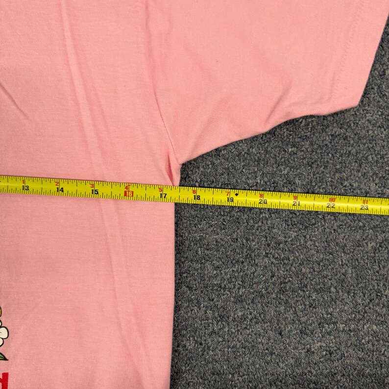 May include: A light pink t-shirt featuring a partial graphic of a flower with white petals and a yellow center. A yellow measuring tape is positioned horizontally across the chest, showing a width measurement of approximately 11 inches.