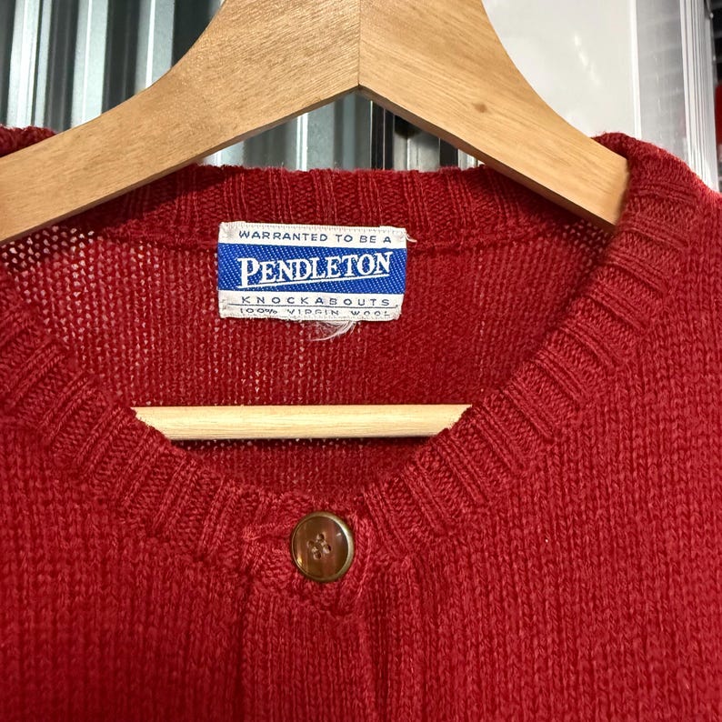 May include: A solid red knit cardigan featuring a ribbed crew neckline and a single brown button closure. A white rectangular label is sewn at the inner collar with blue text reading WARRANTED TO BE A PENDLETON KNOCKABOUTS 100% VIRGIN WOOL.