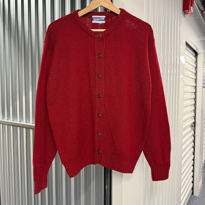 May include: A solid red long-sleeve cardigan sweater featuring a crew neckline, a front button-down closure with brown buttons, and ribbed cuffs and hem. A Pendleton brand label is stitched inside the collar of this knit garment.