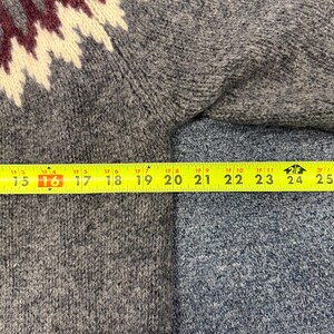 May include: A grey knit sweater featuring a maroon and cream zigzag pattern across the shoulder area. A yellow measuring tape is positioned horizontally across the chest, showing a width measurement of approximately 10 inches from the seam.