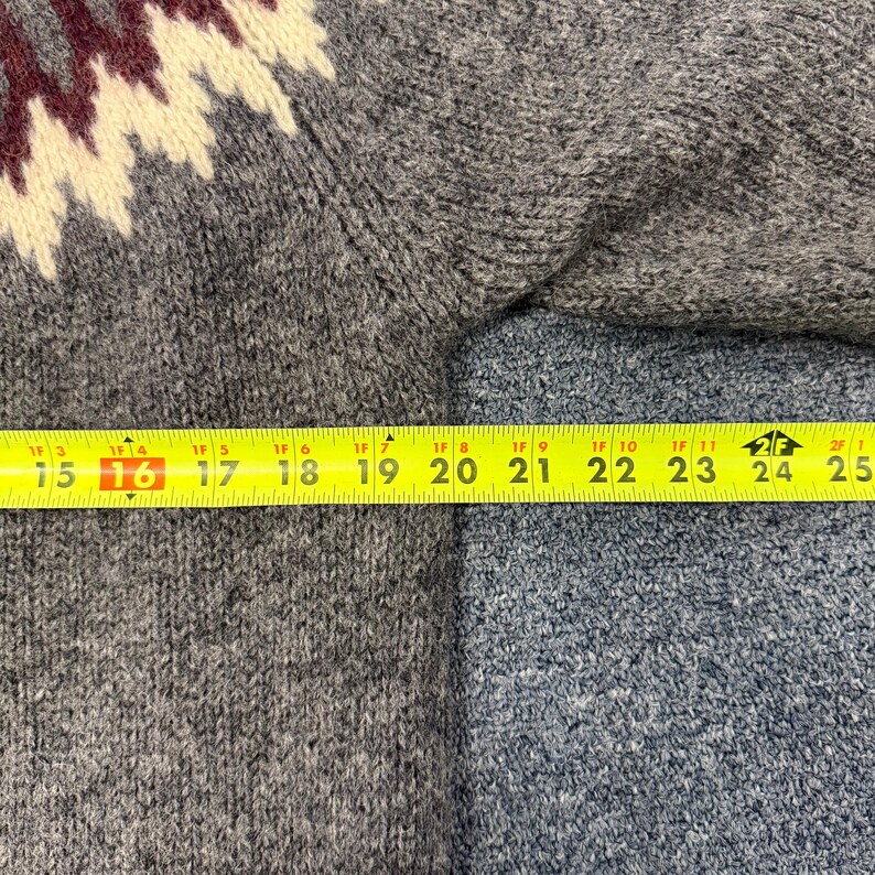 May include: A grey knit sweater featuring a maroon and cream zigzag pattern across the shoulder area. A yellow measuring tape is positioned horizontally across the chest, showing a width measurement of approximately 10 inches from the seam.