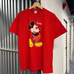 Puede incluir: Una camiseta roja de manga corta con un gr&aacute;fico de Mickey Mouse con sus caracter&iacute;sticos pantalones cortos rojos y zapatos amarillos. La palabra Florida est&aacute; impresa en letra blanca sobre el personaje y se ve una etiqueta en el cuello.