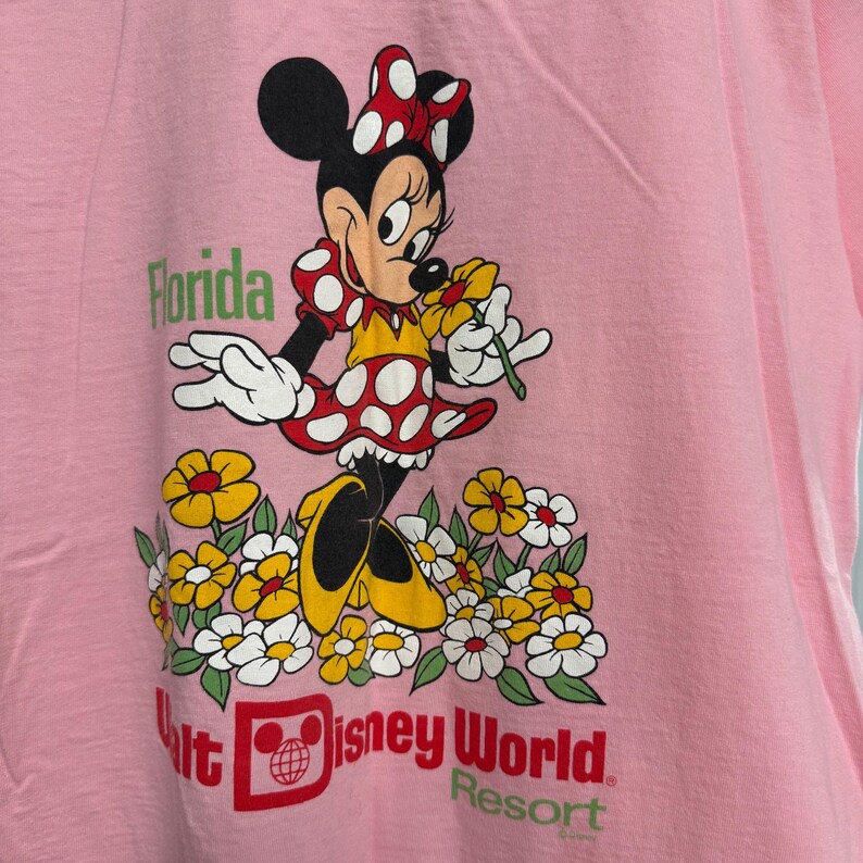 May include: A light pink t-shirt featuring a graphic of Minnie Mouse wearing a red and white polka dot dress and bow, standing in a field of yellow and white flowers. The text reads Florida and Walt Disney World Resort in red and green.