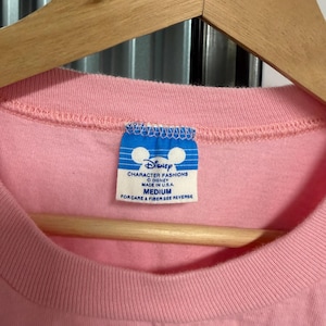 May include: A light pink t-shirt featuring a blue and white rectangular tag at the inner neckline. The tag displays the Disney logo, the text Disney Character Fashions, Made in U.S.A., and the size Medium, with instructions to see the reverse.