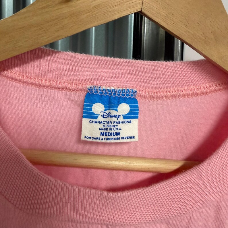 May include: A light pink t-shirt featuring a blue and white rectangular tag at the inner neckline. The tag displays the Disney logo, the text Disney Character Fashions, Made in U.S.A., and the size Medium, with instructions to see the reverse.