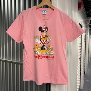 May include: A light pink short-sleeve t-shirt featuring a graphic of Minnie Mouse standing in a field of yellow and white flowers. The text reads Florida and Walt Disney World Resort. The shirt has a crew neckline and is displayed on a wooden hanger.