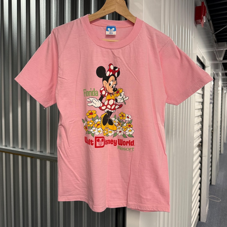 May include: A light pink short-sleeve t-shirt featuring a graphic of Minnie Mouse standing in a field of yellow and white flowers. The text reads Florida and Walt Disney World Resort. The shirt has a crew neckline and is displayed on a wooden hanger.