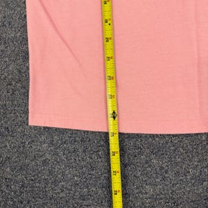 May include: A plain light pink t-shirt featuring a hemmed bottom edge. A yellow measuring tape is placed vertically over the fabric, showing a length measurement extending from 18 inches to 28 inches, indicating a total length of 10 inches.