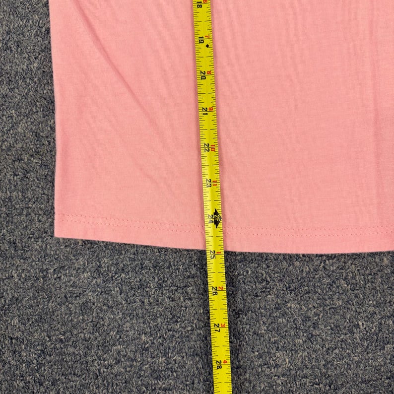 May include: A plain light pink t-shirt featuring a hemmed bottom edge. A yellow measuring tape is placed vertically over the fabric, showing a length measurement extending from 18 inches to 28 inches, indicating a total length of 10 inches.