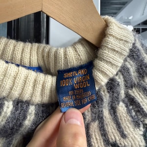 May include: A knit sweater featuring a cream-colored ribbed collar and a dark gray and cream patterned body. A blue fabric tag is attached to the inner collar, printed with the text SHETLAND 100% VIRGIN WOOL, RN 29685, MADE IN MAURITIUS, REVERSE SIDE FOR CARE.