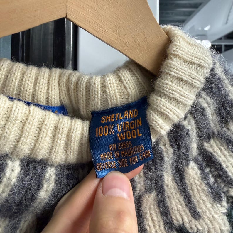 May include: A knit sweater featuring a cream-colored ribbed collar and a dark gray and cream patterned body. A blue fabric tag is attached to the inner collar, printed with the text SHETLAND 100% VIRGIN WOOL, RN 29685, MADE IN MAURITIUS, REVERSE SIDE FOR CARE.