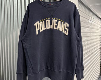 May include: A navy blue long-sleeve crewneck sweatshirt featuring the text RALPH LAUREN POLO JEANS embroidered in a large, arched, cream-colored font with yellow accents across the chest. The garment has a ribbed collar, cuffs, and hem.