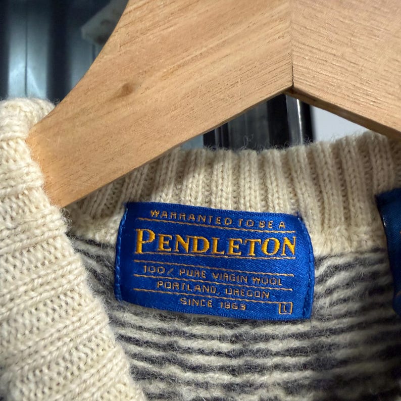 May include: A cream-colored knit sweater featuring a blue rectangular fabric label at the inner neckline. The label reads: WARRANTED TO BE A PENDLETON, 100% PURE VIRGIN WOOL, PORTLAND, OREGON, SINCE 1863, with the size L printed in the bottom right corner.
