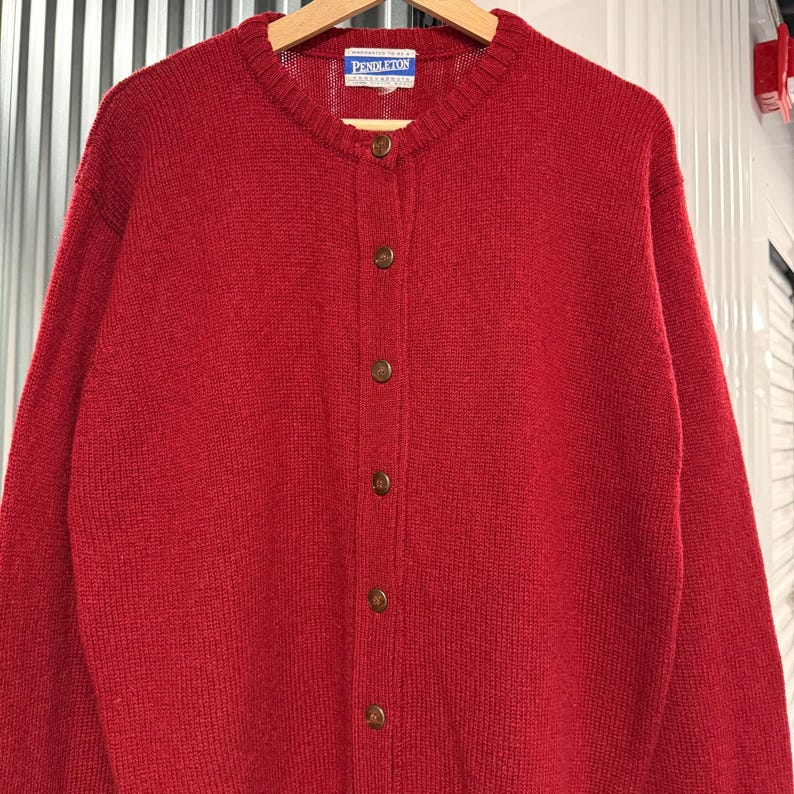 May include: A solid red long-sleeve knit cardigan featuring a crew neckline and a front button closure with five brown buttons. The interior collar tag displays the brand name Pendleton in white text on a blue background, centered at the neck.