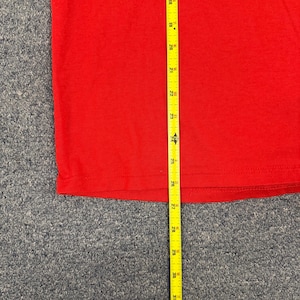 Puede incluir: Una camiseta roja lisa con una cinta m&eacute;trica amarilla colocada verticalmente sobre la tela. La cinta indica una medida de longitud que comienza en 46 cm y se extiende hasta 79 cm, mostrando el dobladillo de la prenda de color rojo.