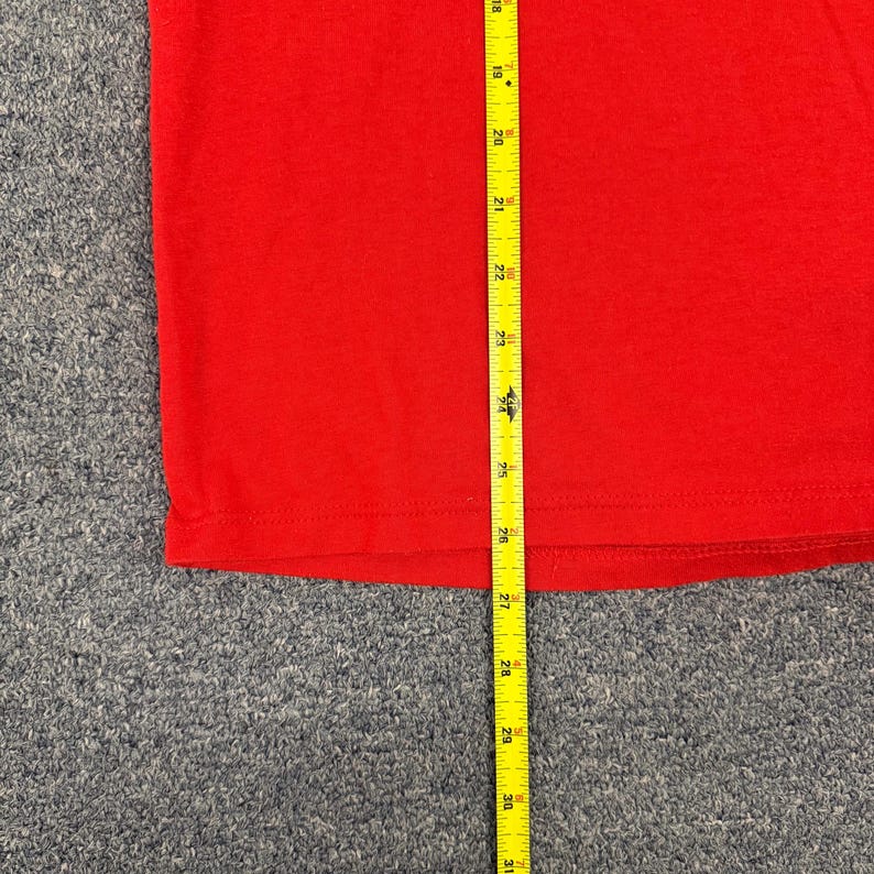 Puede incluir: Una camiseta roja lisa con una cinta m&eacute;trica amarilla colocada verticalmente sobre la tela. La cinta indica una medida de longitud que comienza en 46 cm y se extiende hasta 79 cm, mostrando el dobladillo de la prenda de color rojo.