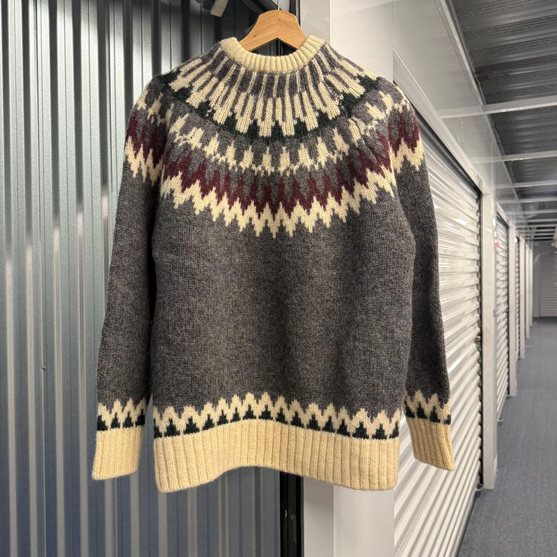 May include: A long-sleeve knit sweater featuring a charcoal gray base with a cream-colored crew neckline, cuffs, and hem. The yoke and hem are decorated with a geometric pattern in shades of cream, dark charcoal, and deep burgundy red.