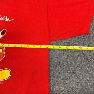 Puede incluir: Una camiseta roja de manga corta con un gr&aacute;fico parcial de un personaje de dibujos animados y la palabra Florida. Una cinta m&eacute;trica amarilla colocada horizontalmente sobre el pecho muestra una anchura de aproximadamente 35,5 cm.