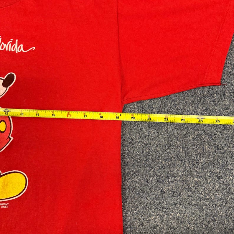 Puede incluir: Una camiseta roja de manga corta con un gr&aacute;fico parcial de un personaje de dibujos animados y la palabra Florida. Una cinta m&eacute;trica amarilla colocada horizontalmente sobre el pecho muestra una anchura de aproximadamente 35,5 cm.