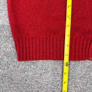 May include: A red knit sweater featuring a ribbed hemline. A yellow measuring tape is positioned vertically against the fabric, showing a length measurement starting from approximately 17.5 inches and extending down to 27 inches.
