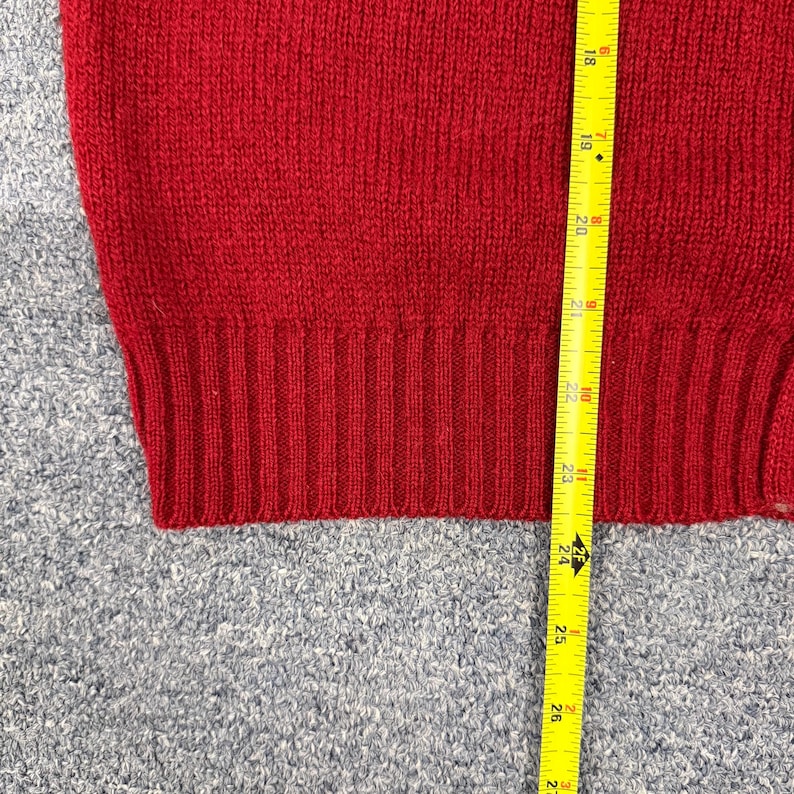 May include: A red knit sweater featuring a ribbed hemline. A yellow measuring tape is positioned vertically against the fabric, showing a length measurement starting from approximately 17.5 inches and extending down to 27 inches.
