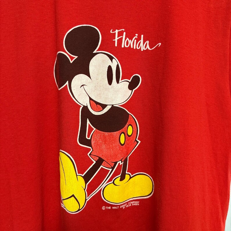 Puede incluir: Una camiseta de color rojo brillante con un estampado gr&aacute;fico de Mickey Mouse de pie. El personaje lleva pantalones cortos rojos con botones amarillos y zapatos amarillos. La palabra Florida aparece en letra cursiva blanca, con el texto The Walt Disney Company.