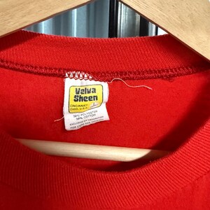 Puede incluir: Una camiseta de color rojo brillante con una etiqueta interior blanca en el cuello con el texto Velva Sheen, Cincinnati, Ohio, U.S.A. La etiqueta indica una talla L y una composici&oacute;n de 50 por ciento poli&eacute;ster y 50 por ciento algod&oacute;n.