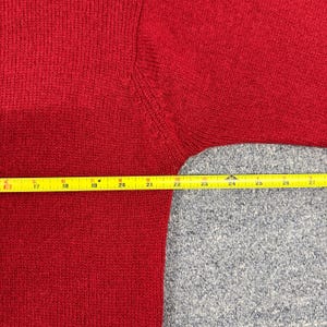 May include: A red knit sweater featuring a visible seam at the shoulder. A yellow measuring tape is positioned horizontally across the garment, showing a measurement span from 16 inches to 27 inches across the chest and sleeve area.
