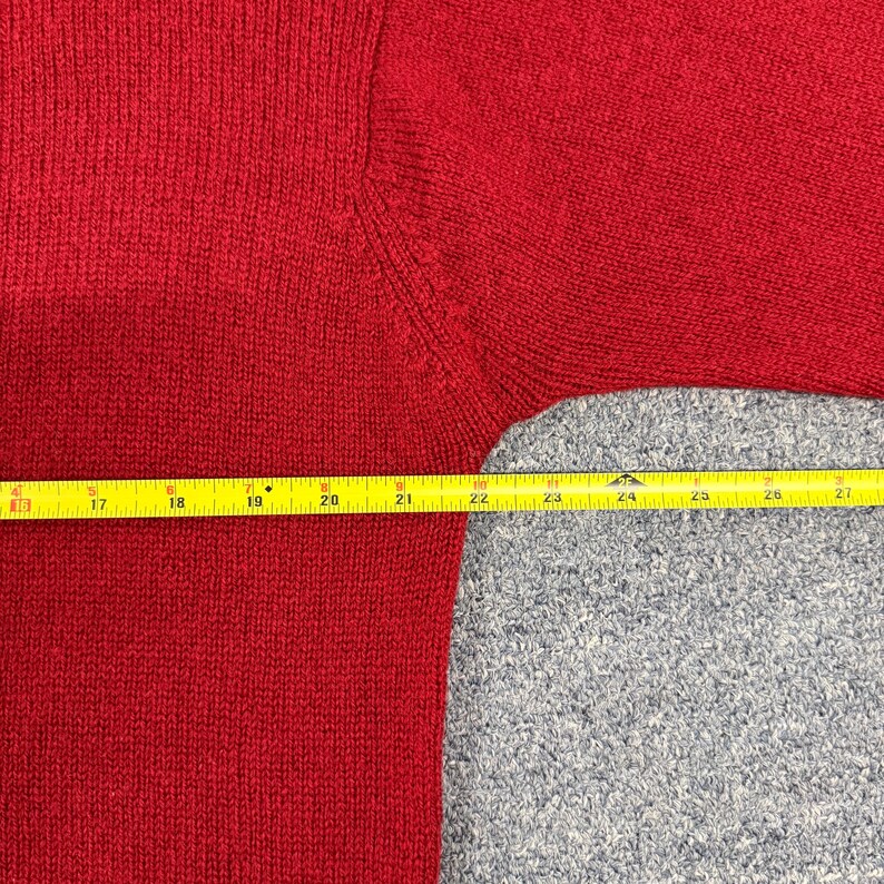 May include: A red knit sweater featuring a visible seam at the shoulder. A yellow measuring tape is positioned horizontally across the garment, showing a measurement span from 16 inches to 27 inches across the chest and sleeve area.