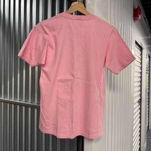 May include: A plain, solid light pink short-sleeve t-shirt with a crew neckline. The garment is displayed hanging on a wooden hanger, showing the back side of the shirt with a smooth, consistent fabric texture and simple hemmed edges.