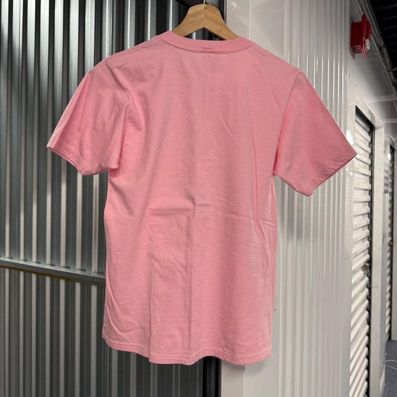 May include: A plain, solid light pink short-sleeve t-shirt with a crew neckline. The garment is displayed hanging on a wooden hanger, showing the back side of the shirt with a smooth, consistent fabric texture and simple hemmed edges.