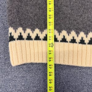 May include: A charcoal gray knit sweater featuring a cream-colored ribbed hem and a cream and black zigzag pattern above the hem. A yellow measuring tape is placed vertically over the garment, showing a length measurement of approximately 12 inches.