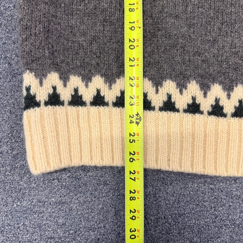 May include: A charcoal gray knit sweater featuring a cream-colored ribbed hem and a cream and black zigzag pattern above the hem. A yellow measuring tape is placed vertically over the garment, showing a length measurement of approximately 12 inches.