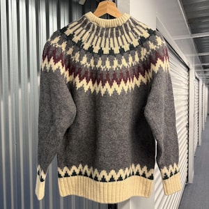 May include: A long-sleeve crew neck sweater featuring a charcoal gray base with a cream, dark green, and burgundy geometric pattern around the yoke and a cream ribbed hem and cuffs. The knit garment has a circular yoke design.