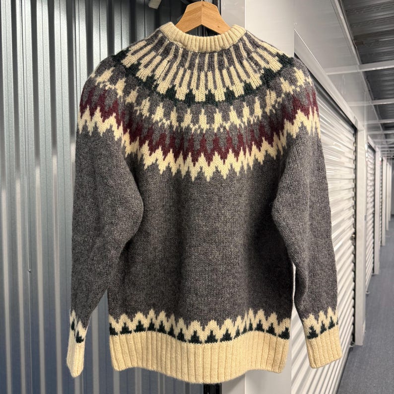 May include: A long-sleeve crew neck sweater featuring a charcoal gray base with a cream, dark green, and burgundy geometric pattern around the yoke and a cream ribbed hem and cuffs. The knit garment has a circular yoke design.