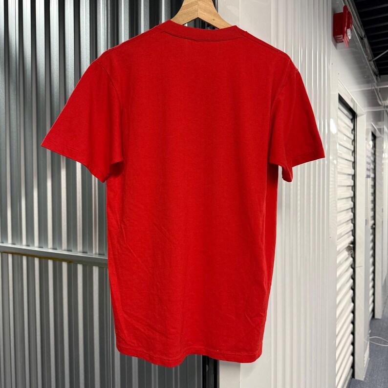 Puede incluir: Una camiseta roja de manga corta con un cuello redondo cl&aacute;sico. La prenda se muestra colgada en una percha de madera, presentando una vista trasera lisa con un dise&ntilde;o sencillo y limpio, manteniendo un color rojo vibrante y uniforme.