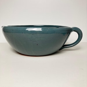 May include: A deep, rounded ceramic bowl with a smooth, glossy teal-blue glaze. The bowl features a single curved handle on the right side, a wide circular opening, and a slightly tapered base, showcasing a solid, uniform color throughout the exterior.