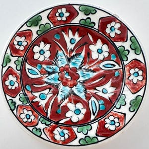 Puede incluir: Un plato de cerámica redondo con un medallón central rojo y un motivo floral azul y blanco. El borde presenta rombos rojos alternados con flores blancas y grupos de hojas verdes, todo delineado en negro sobre una base de color blanco.