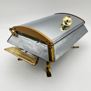 Puede incluir: Un plato de frotamiento rectangular de cromo con acabado plateado pulido, acentuado con adornos de metal dorado, un pomo dorado redondeado en la tapa y cuatro patas doradas curvas. La unidad incluye un asa lateral dorada y un mecanismo de tapa.