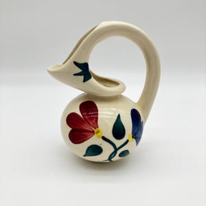 May include: A cream-colored ceramic pitcher with a rounded body and a high, arched handle. The surface is decorated with hand-painted floral motifs featuring red, blue, and green petals and leaves. A small dark blue bird is painted near the spout.
