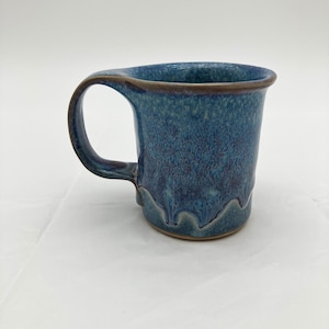 May include: A ceramic mug featuring a mottled blue and purple glaze with a glossy finish. The mug has a wide, curved handle and a flared rim, with a distinct drip pattern near the base that reveals a subtle brown clay tone at the bottom edge.