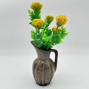 May include: A ceramic pitcher with a brown and grey mottled glaze and a vertical ribbed texture, featuring a handle and a flared rim. The pitcher contains a bouquet of artificial yellow flowers with textured, rounded heads and green foliage.
