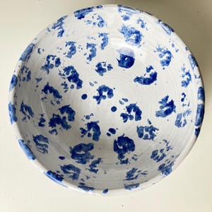 May include: A round ceramic bowl with a white base and a hand-painted blue speckled pattern. The interior and exterior surfaces feature irregular, abstract blue spots of varying sizes distributed across the white glaze, creating a spotted design.