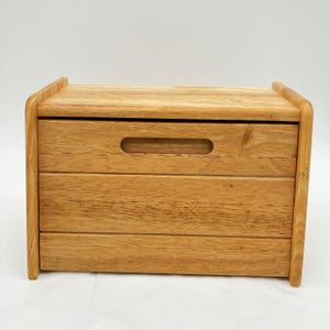 May include: A rectangular wooden bread bin with a light brown natural wood grain finish. The front panel features a horizontal cutout handle centred near the top, and the structure is composed of three distinct horizontal wooden planks.