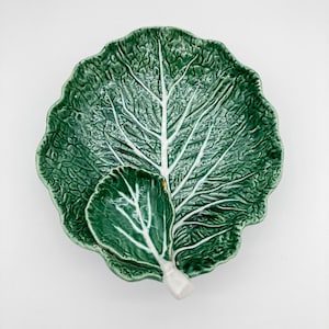 May include: A ceramic dish shaped like a large, textured green cabbage leaf with prominent white veins. A smaller, matching cabbage leaf-shaped dish is attached to the surface, creating a layered, organic appearance with a glossy finish.