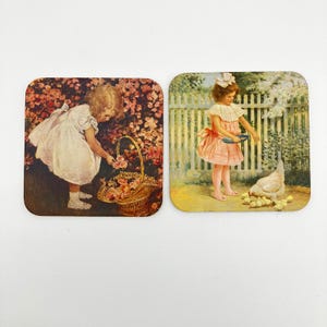 May include: Two rectangular coasters featuring vintage-style illustrations. The left coaster shows a child in a white dress gathering flowers in a basket. The right coaster depicts a child in a pink dress feeding a hen and chicks near a fence.