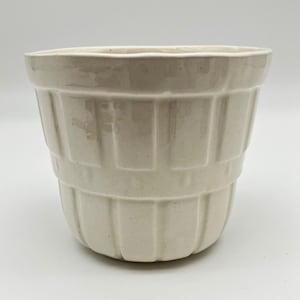 May include: A cream-coloured ceramic planter with a glossy finish, featuring a textured exterior design of vertical panels that mimic the appearance of a wooden basket, with a wide, open top and a tapered base structure.