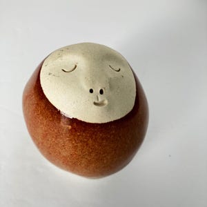 May include: A round ceramic sculpture featuring a smooth, matte off-white face with closed eyes and a small mouth set into a glossy, speckled reddish-brown body. The object has a rounded, pebble-like shape with a distinct contrast between the textured face and the glazed, reflective lower portion of the piece.