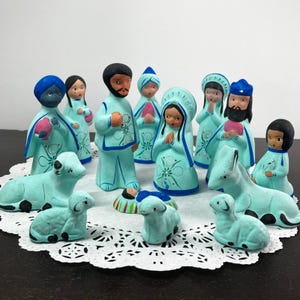 May include: A nativity set featuring eleven hand-painted ceramic figurines in light blue with dark blue and green accents. The set includes human figures, a baby in a manger, and various animals, all arranged on a white paper doily.