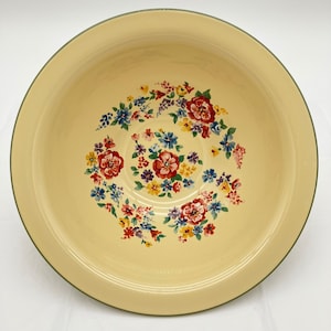 Op de afbeelding: Een ronde, crèmekleurige emaille kom met een brede rand en een centraal bloemmotief. Het interieurontwerp bestaat uit trossen rode, blauwe, gele en paarse bloemen met groene bladeren die in een cirkelvormig patroon op de bodem zijn gerangschikt.